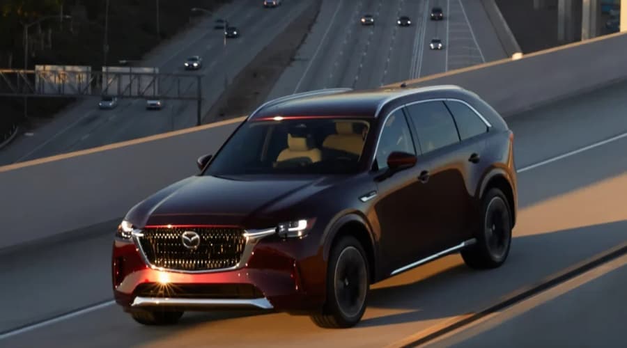Dark red 2026 Mazda CX-90 three-row SUV with i-Activ AWD driving along an elevated highway overpass near Ramsey, NJ during a late afternoon autumn sunset.