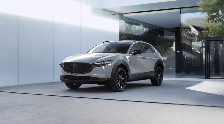 Front three-quarter view of a light grey 2026 Mazda CX-30 parked outside a modern architectural building, showcasing black alloy wheels and sleek crossover body styling for Ramsey and Bergen County, NJ drivers.