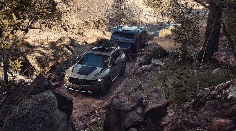 A 2026 Mazda CX-50 Turbo with a loaded roof rack towing a compact teardrop trailer on a rocky off-road trail, highlighting its 3,500-pound towing capacity