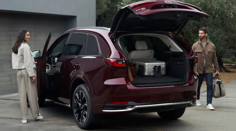 Open power liftgate of an Artisan Red 2026 Mazda CX-90 showing spacious rear cargo space as a couple loads suitcases for an autumn weekend road trip in Ramsey, NJ.