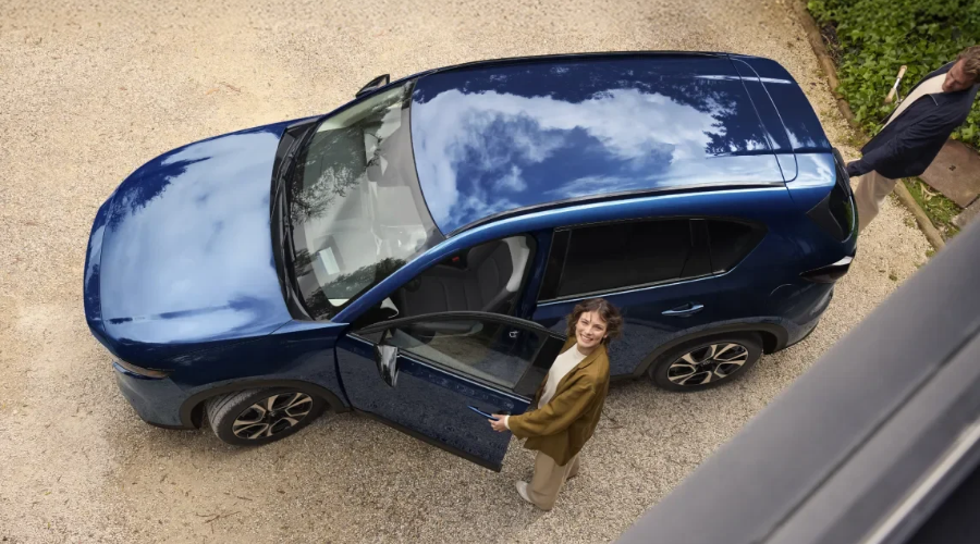 High-angle overhead view of a Deep Crystal Blue Mica 2026 Mazda CX-5 crossover on a gravel driveway with a driver opening the door, highlighting windshield wiper sensor coverage and i-Activ AWD alloy wheels serviced at Ramsey Mazda in Bergen County, NJ