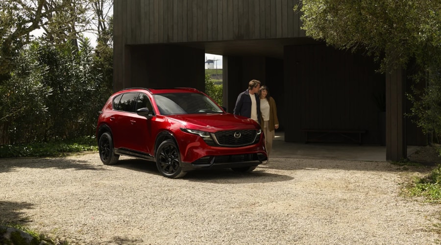 Red 2026 Mazda CX-5 parked on a gravel driveway outside a modern suburban Ramsey, NJ home, as a couple walks toward the crossover before an autumn commute in Bergen County