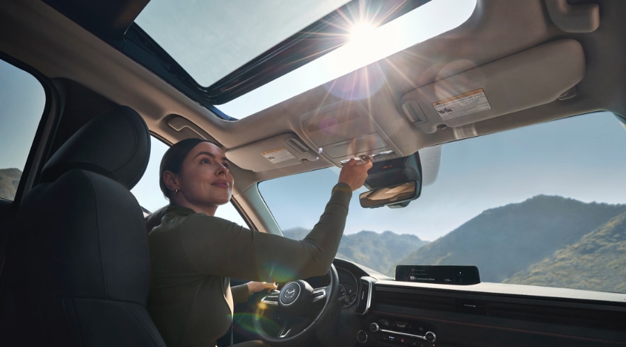 Driver adjusting the panoramic moonroof controls in a 2026 Mazda CX-50 Hybrid interior while commuting through scenic Bergen County, New Jersey.