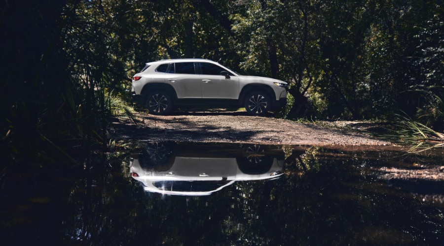 White 2026 Mazda CX-50 Hybrid reflecting in a water puddle on a shaded dirt path in Mahwah, New Jersey, highlighting Mi-Drive Trail Mode capability