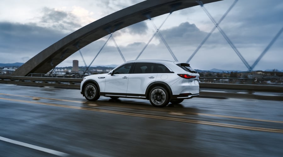 Side rear profile of a white 2026 Mazda CX-90 SUV driving on a wet bridge highway during rainy weather in Bergen County, NJ, demonstrating radar cruise control and safety features.