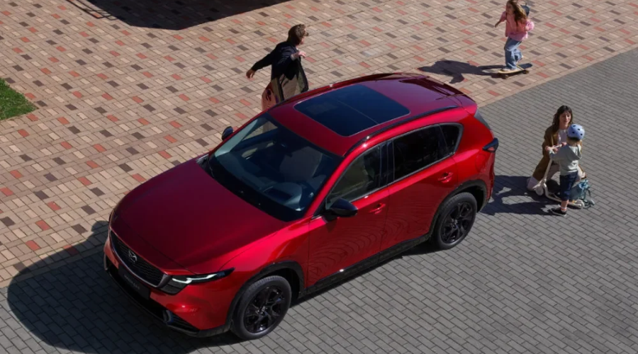 High-angle view of a Soul Red Crystal Metallic 2026 Mazda CX-5 crossover parked on a paved driveway with a family, highlighting the front emblem radar badge and windshield camera sensors for i-Activesense safety tech at Ramsey Mazda in Bergen County, NJ