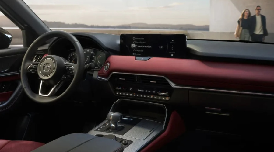 Driver's cockpit view of the 2026 Mazda CX-70 dashboard featuring a leather-wrapped steering wheel, red accents, digital display, and dual-zone climate controls during a morning drive through Bergen County, NJ.