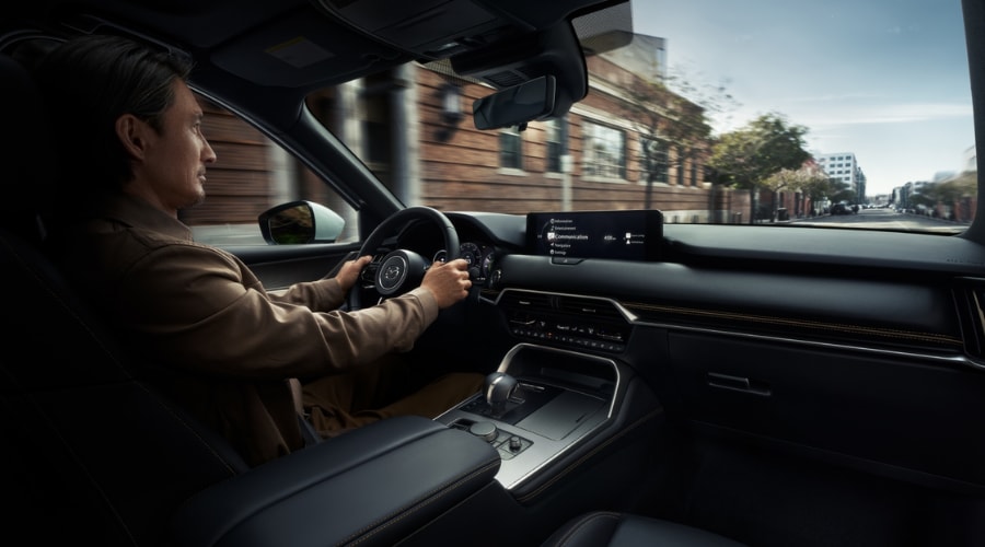 Man driving a 2026 Mazda CX-90 three-row SUV through suburban streets in Bergen County, NJ, utilizing the Active Driving Display and ergonomic safety controls.