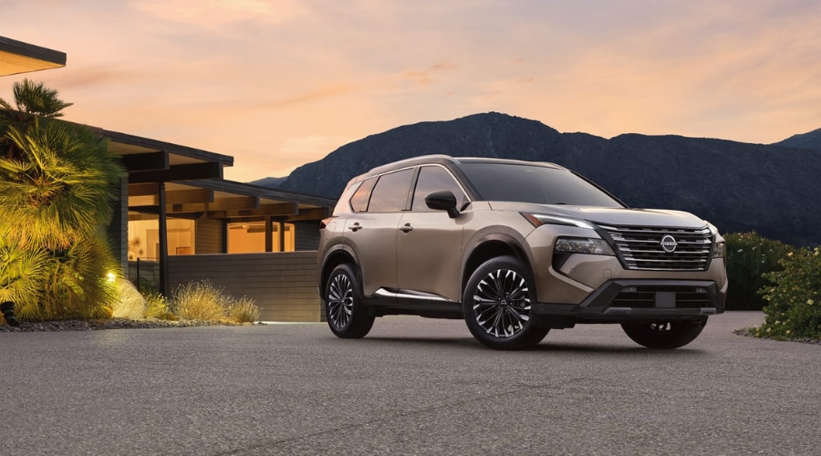 Front quarter view of a champagne gold 2026 Nissan Rogue parked in a driveway at sunset, showcasing the front V-motion grille radar emblem and camera sensors available at Ramsey Nissan in Bergen County, NJ.