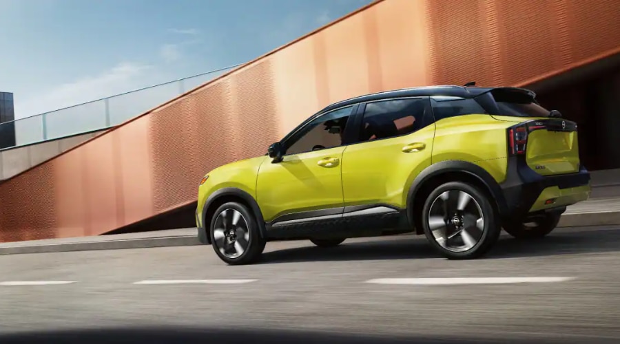 Rear three-quarter profile of a two-tone yellow and black 2026 Nissan Kicks driving past a modern bronze architectural wall, highlighting its athletic high ground clearance stance