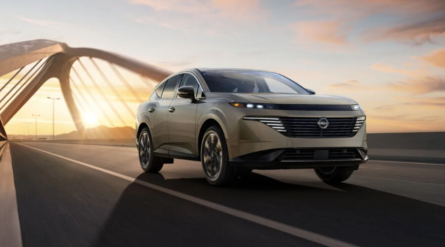 Front three-quarter view of a champagne gold 2026 Nissan Murano driving across a modern arched highway bridge at sunset, ideal for Route 17 and Route 4 commuters in Bergen County, NJ.