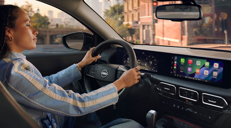 Side view of a driver in a 2026 Nissan Sentra cabin with hands on the leather-wrapped steering wheel, showcasing active Apple CarPlay maps on the widescreen display and digital instrument panel.