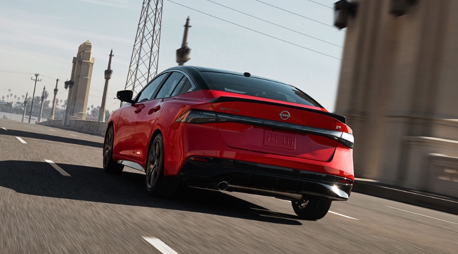 Dynamic rear view of a two-tone red and black 2025 Nissan Sentra SR driving across a bridge, showing the sleek LED taillights, rear spoiler, and lower bumper sonar nodes.