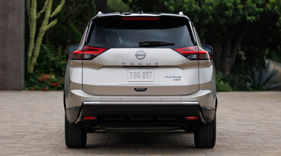 Direct rear view of a champagne gold 2026 Nissan Rogue Platinum AWD showing the rear emblem, Platinum badge, and bumper sonar sensors for Safety Shield 360 at Ramsey Nissan in Bergen County, NJ. 