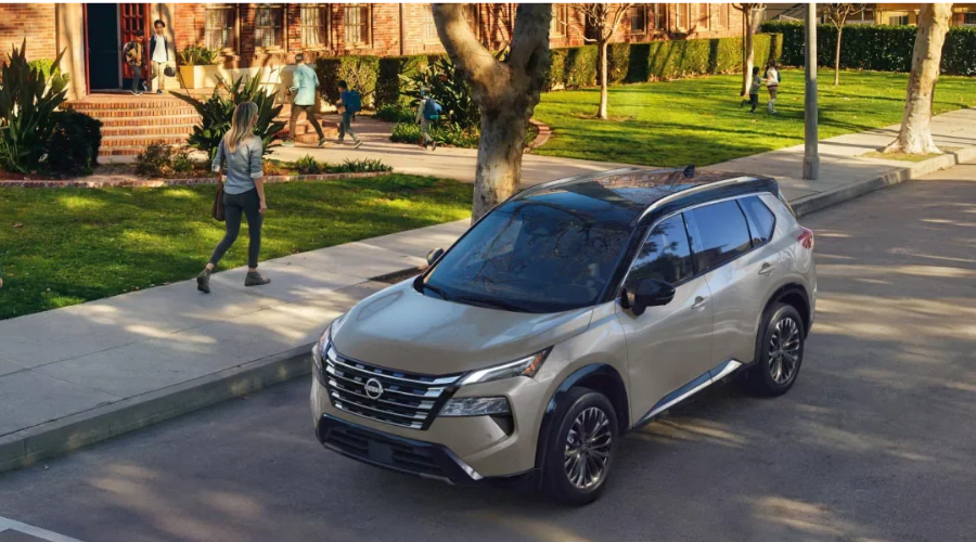 Elevated front-three-quarter view of a champagne metallic 2027 Nissan Rogue SV with a gloss-black panoramic roof and multi-spoke alloy wheels parked along a tree-lined residential street near Bergen County, NJ.
