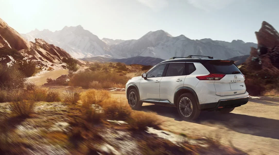 Glacier White 2027 Nissan Rogue SV SUV with a black contrast roof, black roof rails, and Intelligent All-Wheel Drive navigating an unpaved scenic mountain road, on display at Ramsey Nissan in Ramsey, NJ.