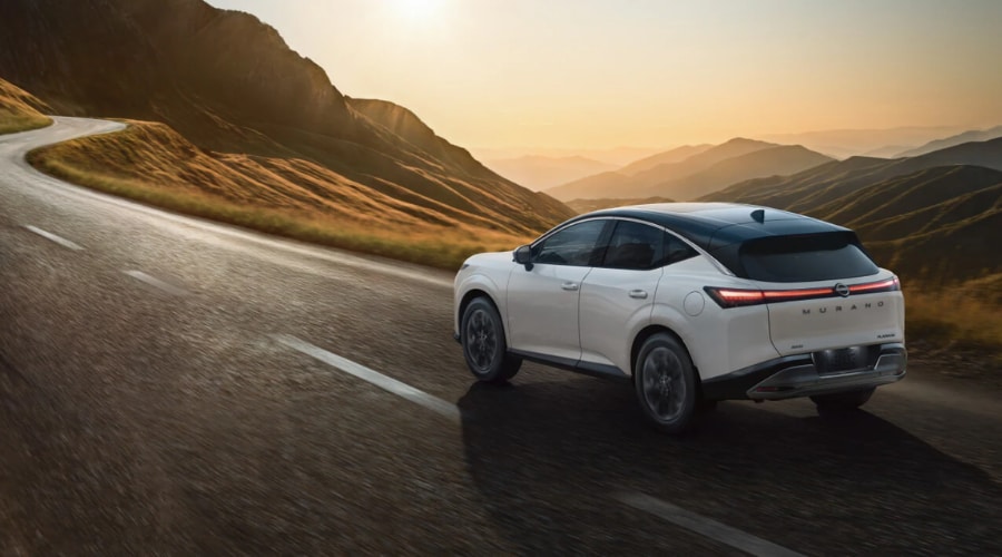 Rear three-quarter view of a two-tone pearl white 2026 Nissan Murano Platinum driving along a winding mountain road at sunset, demonstrating all-weather handling for Bergen County and Rockland drivers.