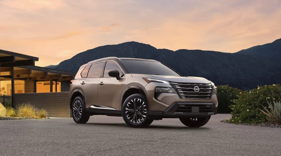 Champagne metallic 2027 Nissan Rogue SV crossover featuring upgraded 18-inch alloy wheels, sleek V-motion grille, and LED headlights parked on a paved driveway at sunset near Ramsey, NJ.