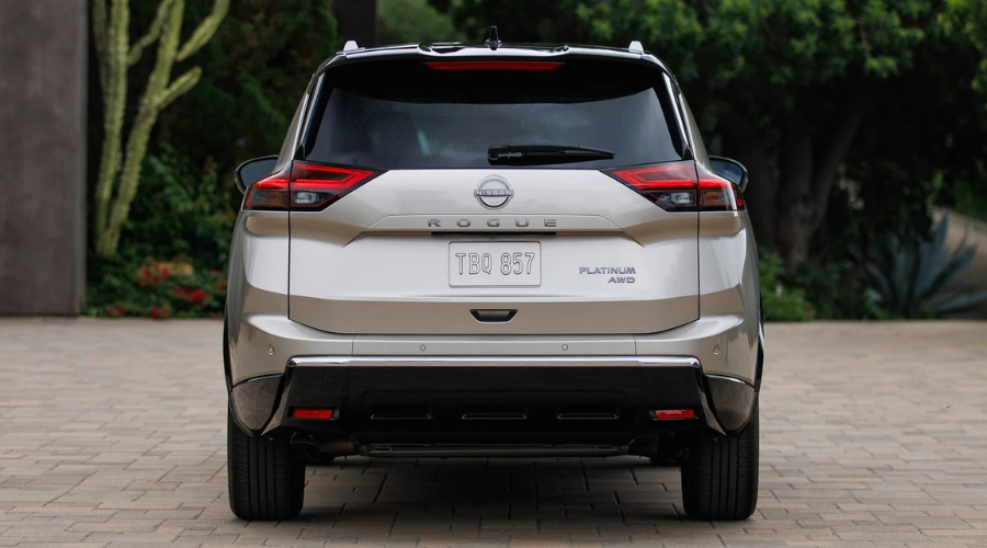 Rear exterior view of a 2027 Nissan Rogue crossover showing the sculpted liftgate with prominent chrome ROGUE badging, rear parking sensors, roof spoiler, and dual-tier LED taillights parked on a paved driveway near Bergen County, NJ.