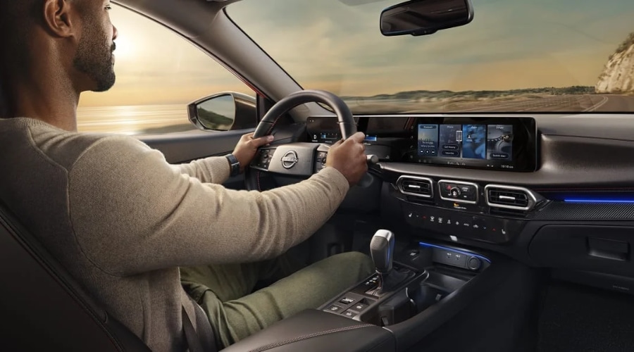 Driver inside the cockpit of a 2027 Nissan Sentra holding the flat-bottom steering wheel, showcasing the dual 12.3-inch digital touchscreen displays and ambient cabin lighting, available for a test drive at Ramsey Nissan in Ramsey, NJ.