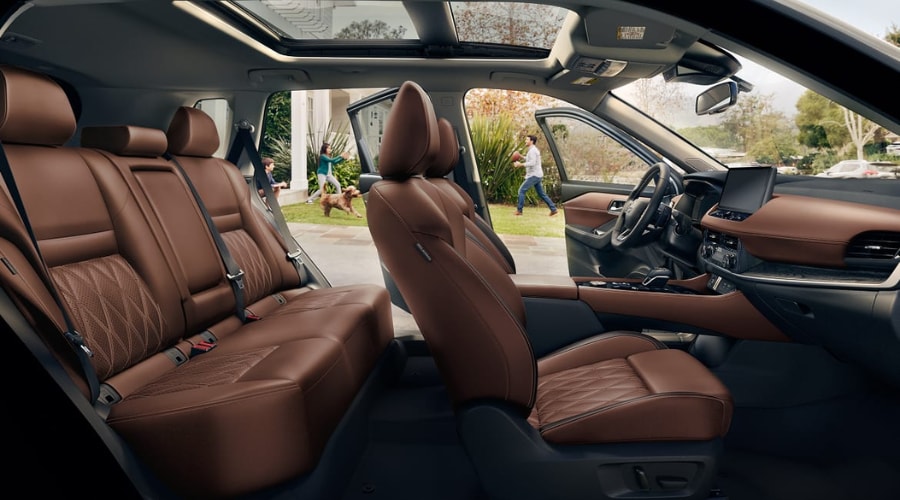 Spacious five passenger 2026 Nissan Rogue interior featuring premium brown quilted leather seats and a panoramic sunroof with doors open to a sunny yard