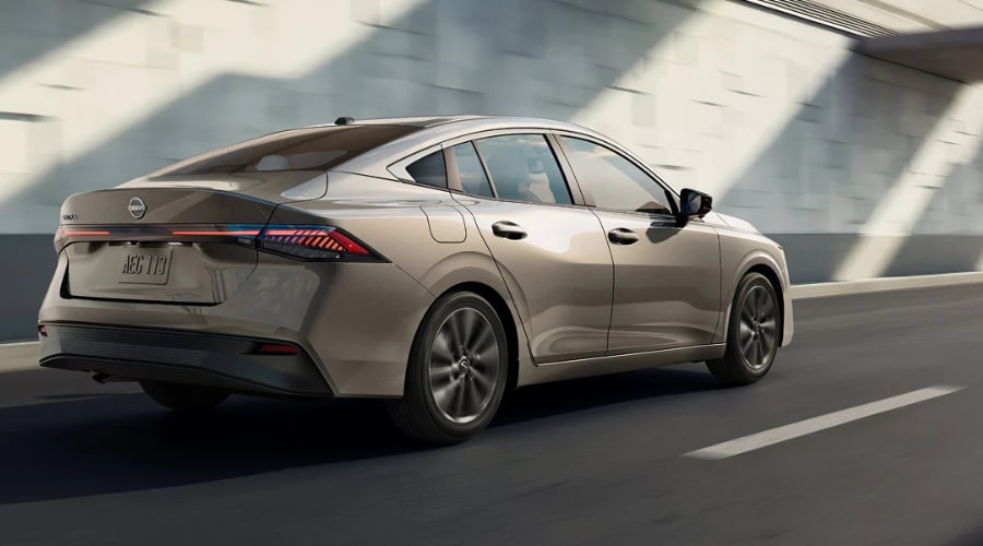 Rear three-quarter view of a champagne metallic 2027 Nissan Sentra sedan driving on an open road, highlighting its sleek fastback silhouette and continuous LED taillight bar, available now at Ramsey Nissan in Ramsey, NJ.