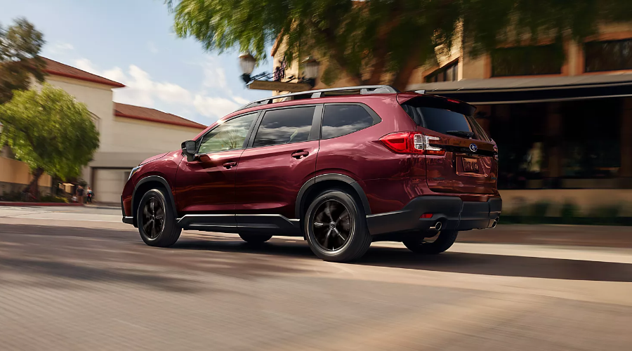 Rear three-quarter action view of a dark red 2027 Subaru Ascent driving through a downtown commercial district in Wyckoff, NJ, showcasing its LED taillights, roof rails, dual exhaust, and black alloy wheels.
