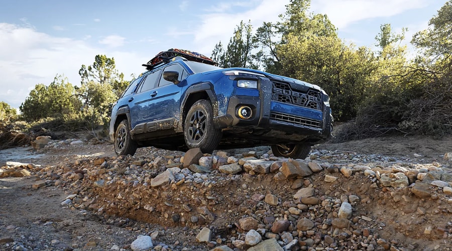 Front three-quarter view of a blue 2026 Subaru Forester driving over a rocky trail with a loaded roof cargo basket, demonstrating X-MODE traction and high ground clearance near Ramsey, NJ.