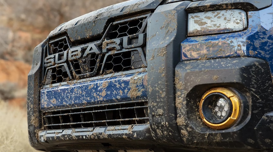 Close-up of a mud-splattered blue Subaru Outback front grille and bumper with gold-ringed fog lights, highlighting the front camera and rugged off-road EyeSight sensor care for North Jersey trail drivers in Mahwah and Ramsey, NJ.