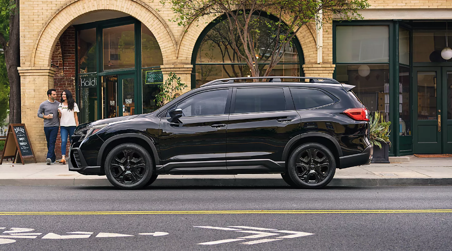 Side profile of a black 2027 Subaru Ascent Onyx Edition parked along a downtown street storefront in Ridgewood, NJ, showing its dark alloy wheels, roof rails, and sleek three-row crossover silhouette.