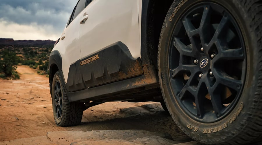 Close-up side view of a white 2026 Subaru Crosstrek driving on a muddy dirt trail, showing black alloy wheels, all-terrain tires, and protective lower body cladding near Ramsey, NJ.