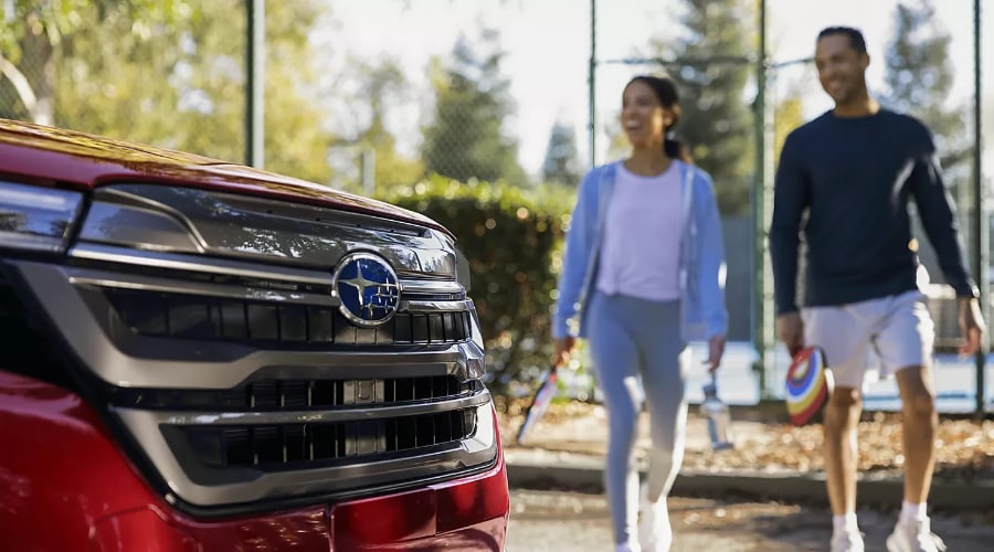 Close-up of the front grille and Subaru star cluster badge on a red 2026 Subaru Forester with a couple walking past at a park in Ramsey, NJ after a morning game of pickleball