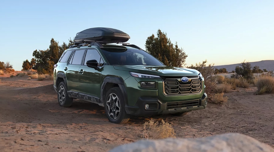 Front three-quarter view of a green Subaru Outback fitted with a roof cargo carrier box, parked on a dirt trail surrounded by desert brush, highlighting its rugged off-road capability for outdoor trips in Bergen County, NJ.