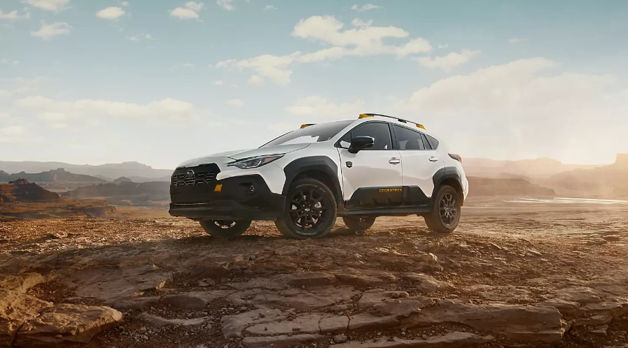 White 2026 Subaru Crosstrek Wilderness compact SUV featuring black lower cladding, gold roof rail accents, and raised ground clearance parked on rugged terrain, highlighting Symmetrical AWD and X-MODE traction for wet weather driving in Ramsey, NJ and Bergen County.