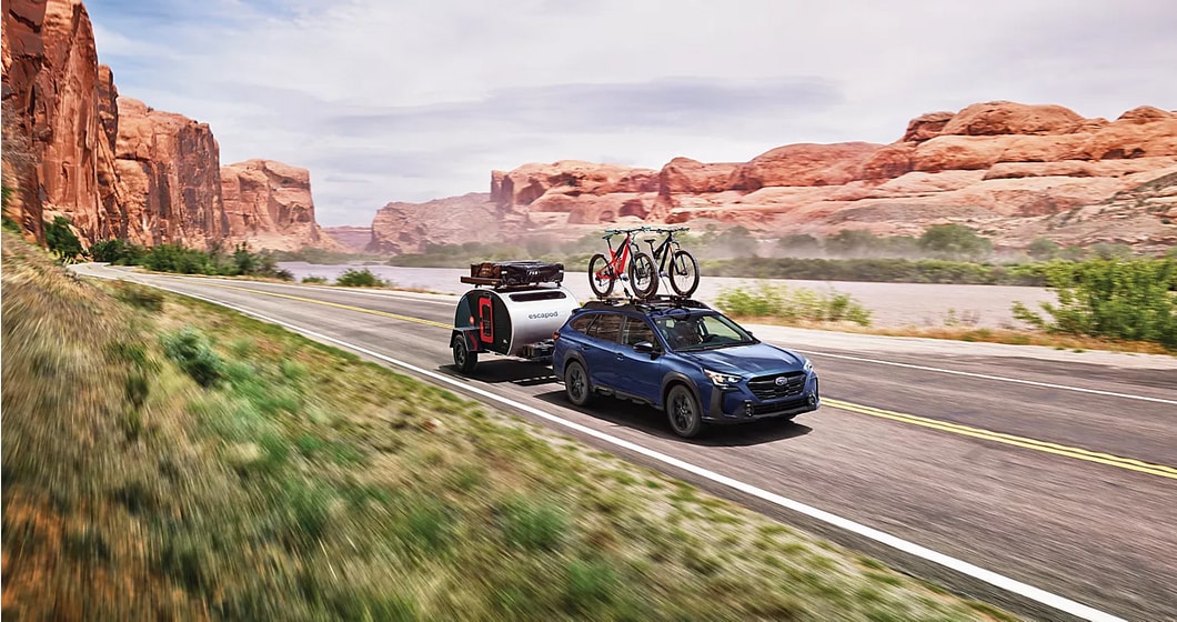 The 2024 Subaru Outback driving on an open road with trees near by two bikes on the back rack hauling cargo