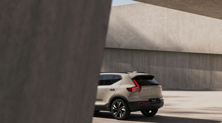 Rear three-quarter view of a beige 2026 Volvo XC40 compact SUV framed by concrete architecture, highlighting illuminated vertical LED taillights and Rear Auto Brake safety tech for Ramsey, NJ drivers.