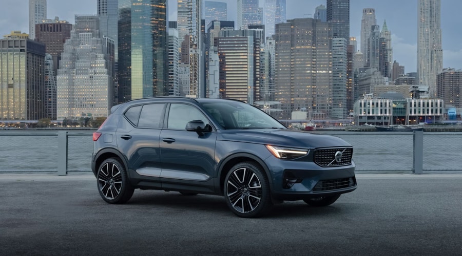 Dark metallic blue 2026 Volvo XC40 compact SUV with active bending pixel LED headlights parked along a waterfront with a city skyline backdrop, representing autumn safety tech for Bergen County and Ramsey, NJ commuters.