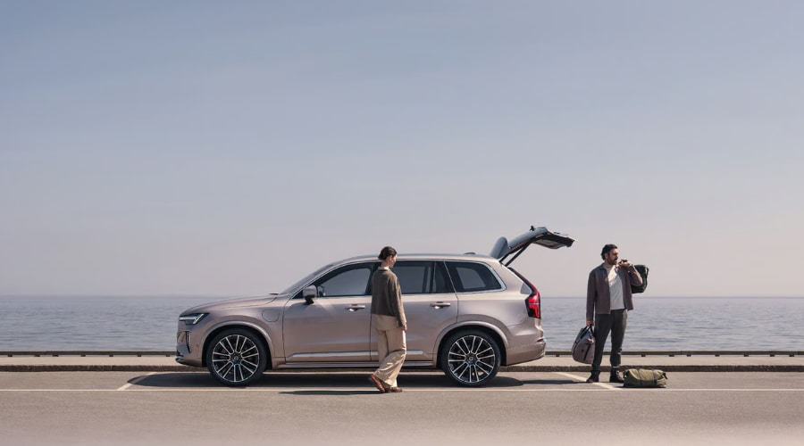 Side profile view of a metallic 2026 Volvo XC90 luxury SUV with its hands-free power tailgate open along a scenic road, showing a Bergen County family loading weekend luggage into the spacious 3rd-row cargo area near Ramsey, NJ.