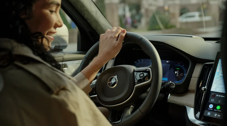 Woman smiling while driving a 2027 Volvo XC90 luxury SUV through a local neighborhood, showcasing the ergonomic leather-wrapped steering wheel, 12.3-inch digital driver display, light wood dash trim, and vertical Google touchscreen navigation, available at Volvo Cars Ramsey near Route 17.