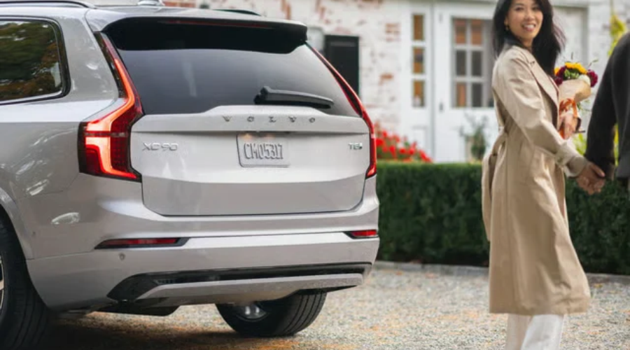 Rear three-quarter view of a silver 2027 Volvo XC90 T8 Plug-In Hybrid luxury SUV parked in a suburban driveway, featuring iconic vertical LED taillights and rear badging, as a stylish woman holding flowers walks towards a residential home in Bergen County, NJ.