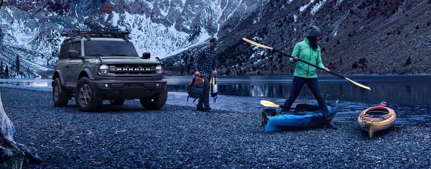 Two people carrying canoes from a grey 2024 Ford Bronco for sale parked near a river.