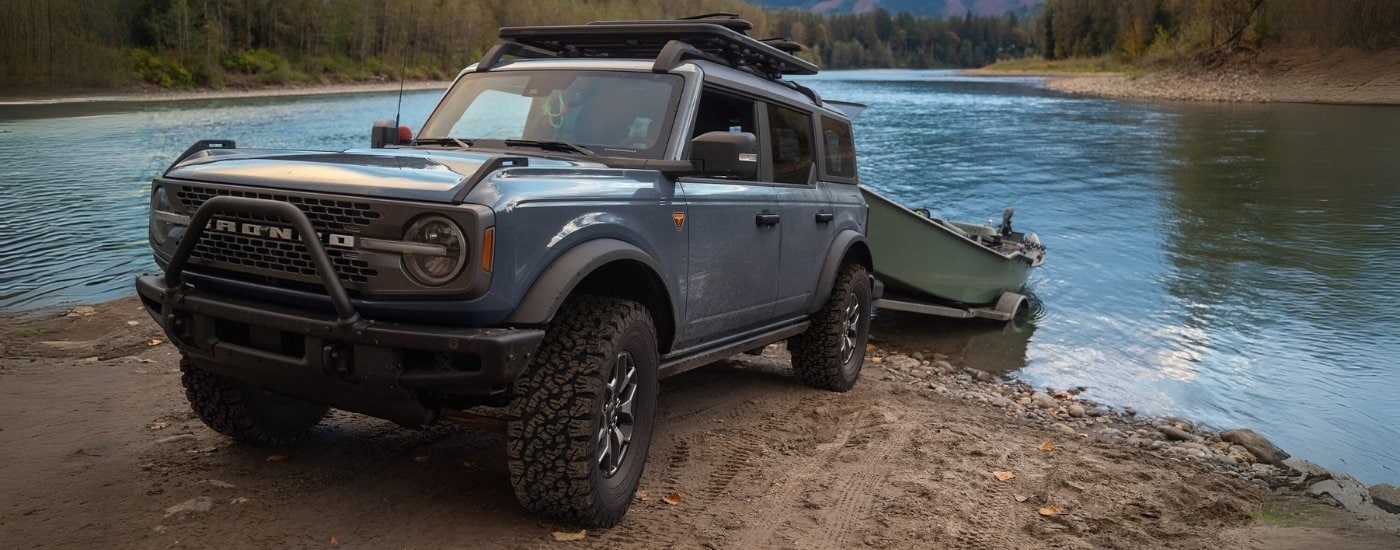 A blue 2025 Ford Bronco Badlands is shown towing a boat near a river.