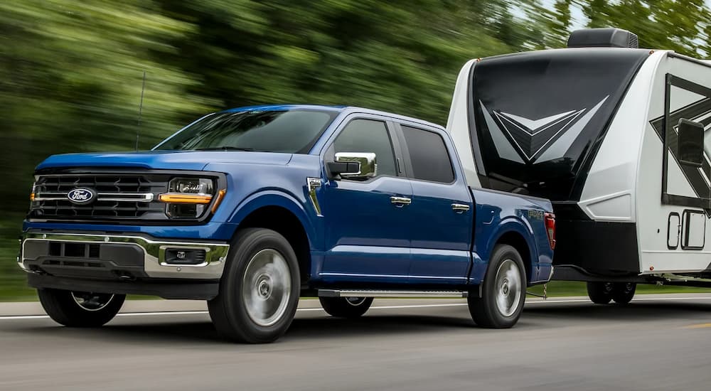 A blue 2024 Ford F-150 XLT is towing a camper on a tree-lined road.