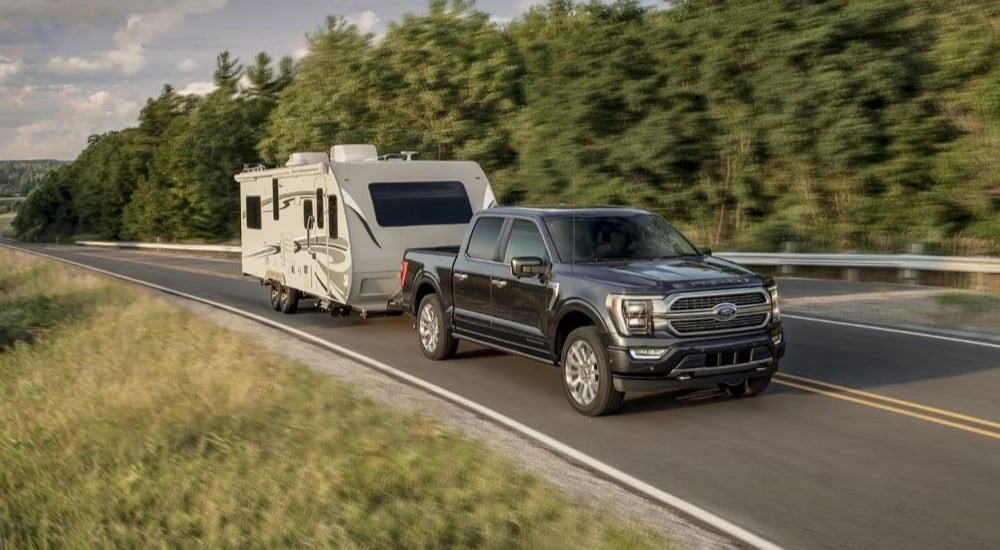 A grey 2023 Ford F-150 Limited is towing a white camper on a tree-lined highway.