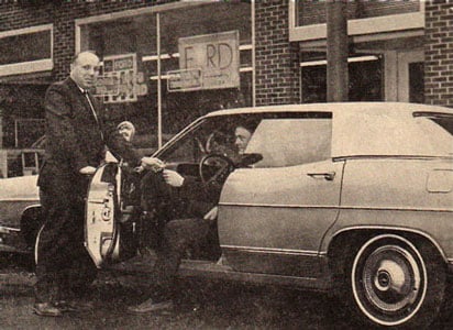Black and white photo of two men with an antique car parked on the street