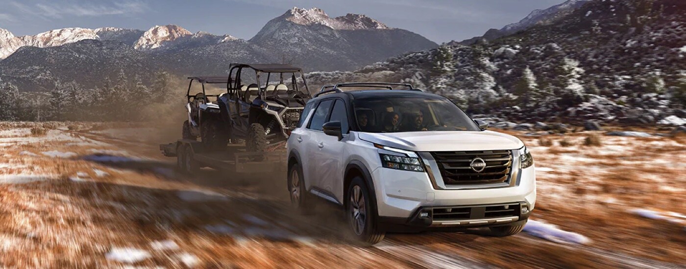 A white 2024 Nissan Pathfinder Platinum towing ATVs in the mountains.
