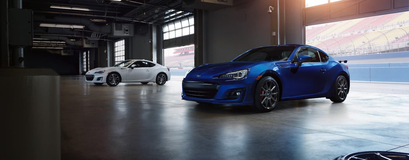 A white and a blue 2022 Subaru BRZ are shown in a large empty garage at a racetrack.