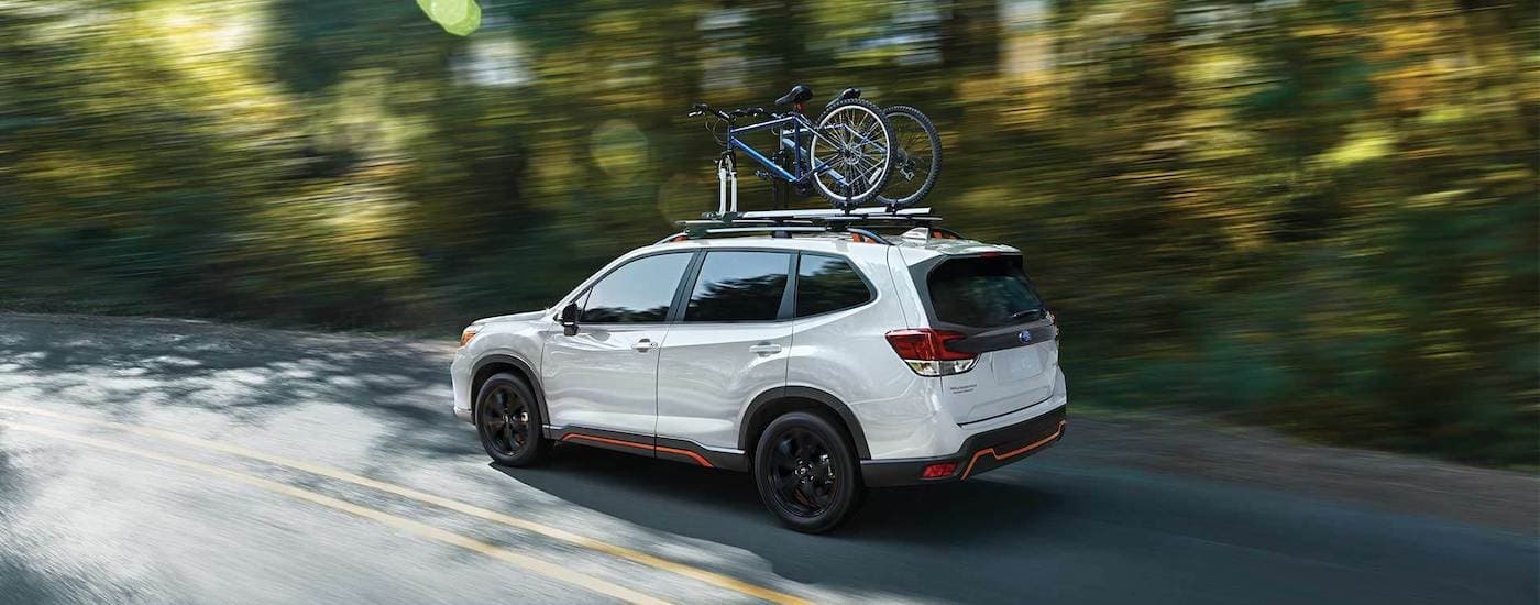 A white 2021 Subaru Foreseter is shown from the side driving past blurred trees with bikes on the roof