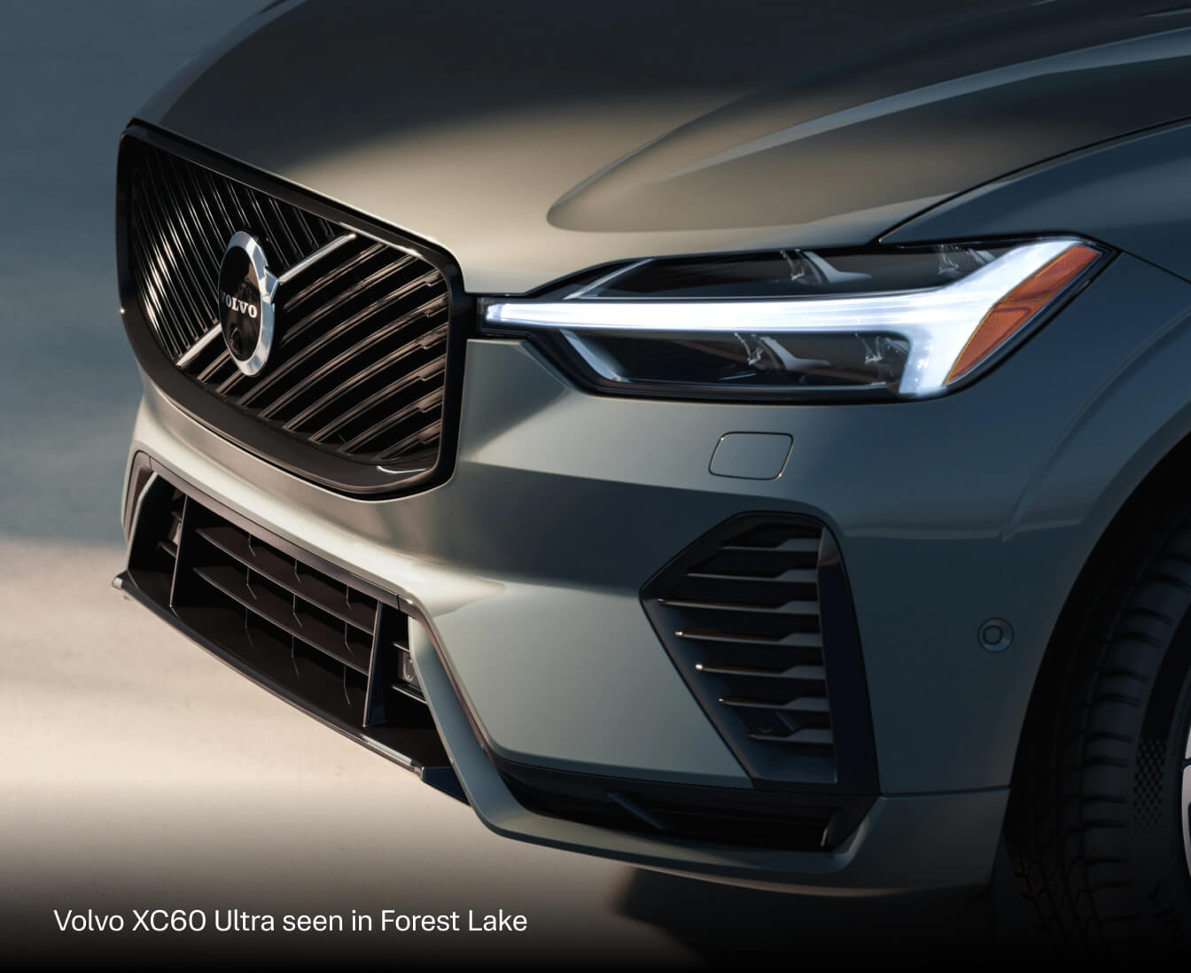 2027 Volvo XC60 Ultra in Forest Lake, close-up of the front grille, Volvo badge and illuminated headlight.