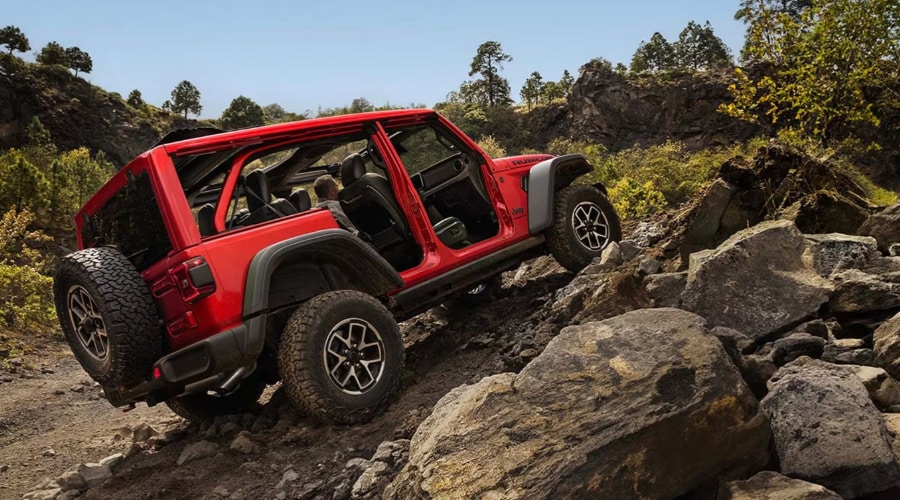 Side rear perspective of a bright red 2026 Jeep Wrangler Rubicon with doors off rock crawling up a steep rocky trail, demonstrating Rock-Trac 4x4 capability and all-terrain tire articulation serviced at Route 18 Autos in East Brunswick, NJ.